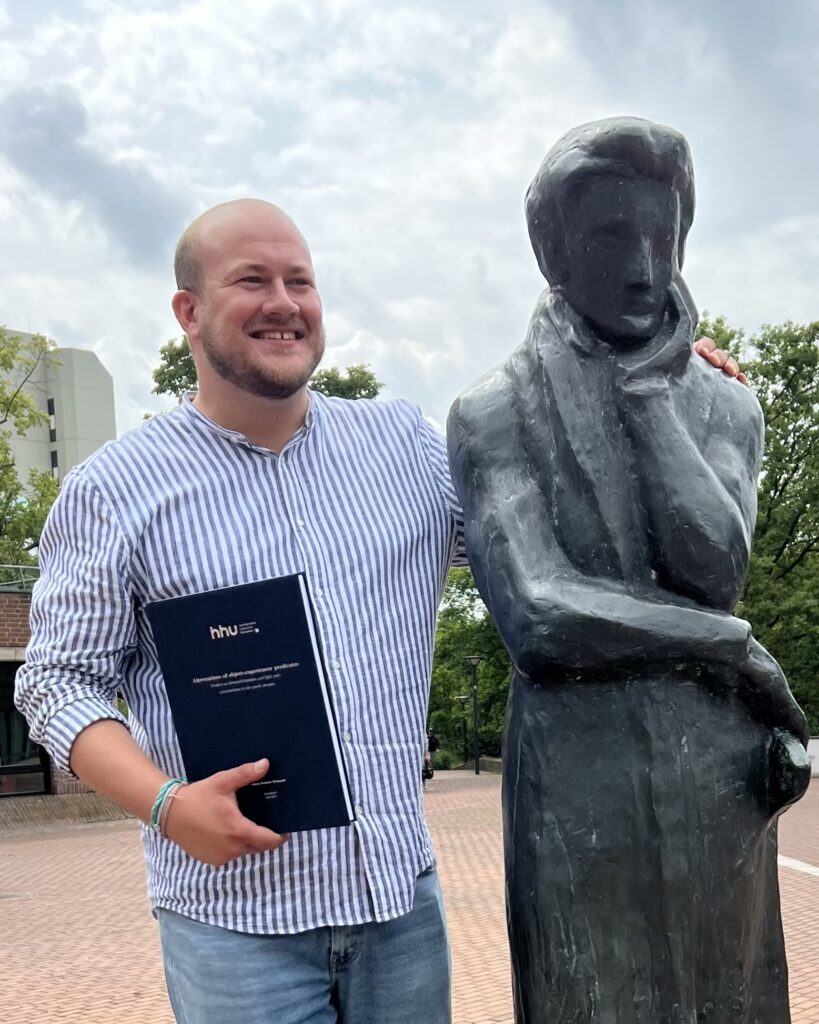 Niklas Wiskandt, with a print copy of his PhD thesis, next to a statue of Heinrich Heine on the campus of HHU Düsseldorf